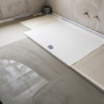 Receveur de douche blanc posé à côté d’une chape ciment neuve en cours de séchage dans une salle de bains en rénovation.