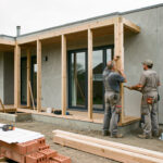 Extension contemporaine en construction accolée à une maison, avec deux artisans, des matériaux et des dessins architecturaux éclairés par la lumière naturelle.
