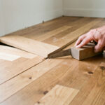 Vue rapprochée de deux parties de parquet en chêne raccordées après l’ouverture d’une cloison, avec des outils de menuiserie et des mains ajustant les lames.